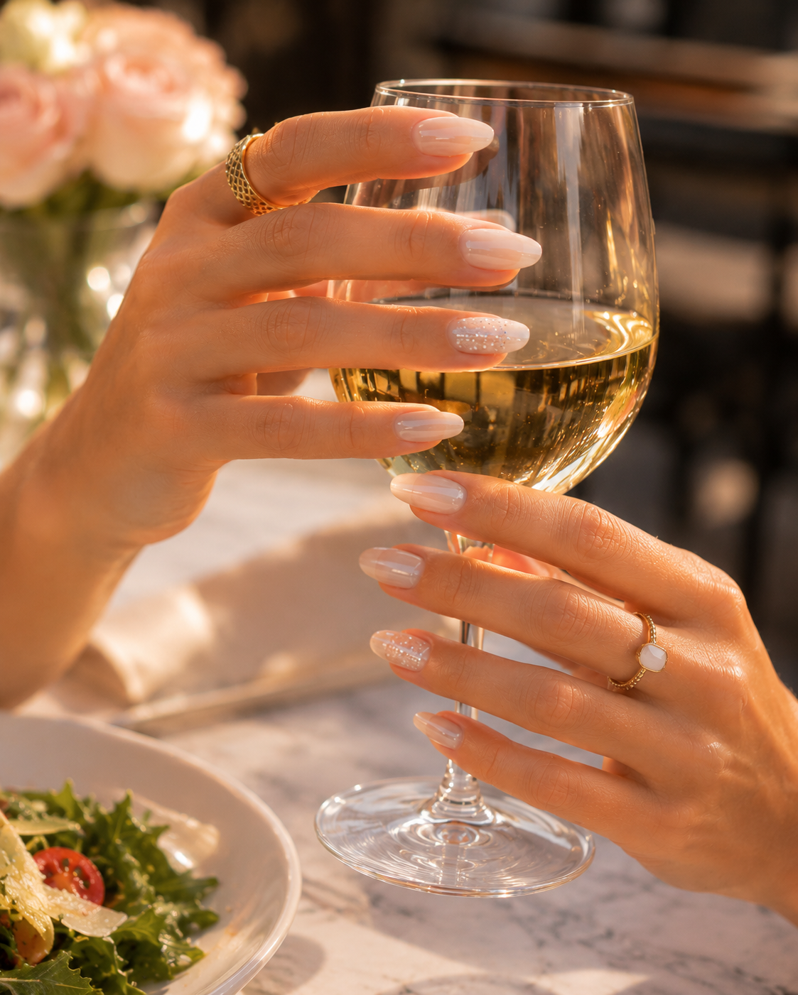 Close-up of nude blush almond-shaped press-on nails with fine glitter accent nails wrapped around a white wine glass in a warm golden-hour café setting.