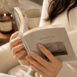 Woman holding an open book while wearing glossy rose chrome press-on nails, styled in a cozy neutral-toned indoor setting with soft candlelight and warm aesthetic decor.