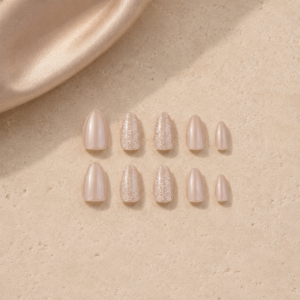 Set of nude blush press-on nails with subtle shimmer and glitter accent nails arranged on a beige stone surface with satin fabric, luxury nail product flat lay.