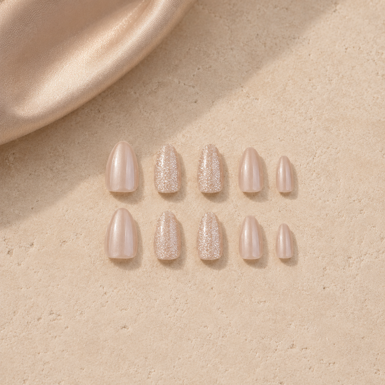Set of nude blush press-on nails with subtle shimmer and glitter accent nails arranged on a beige stone surface with satin fabric, luxury nail product flat lay.