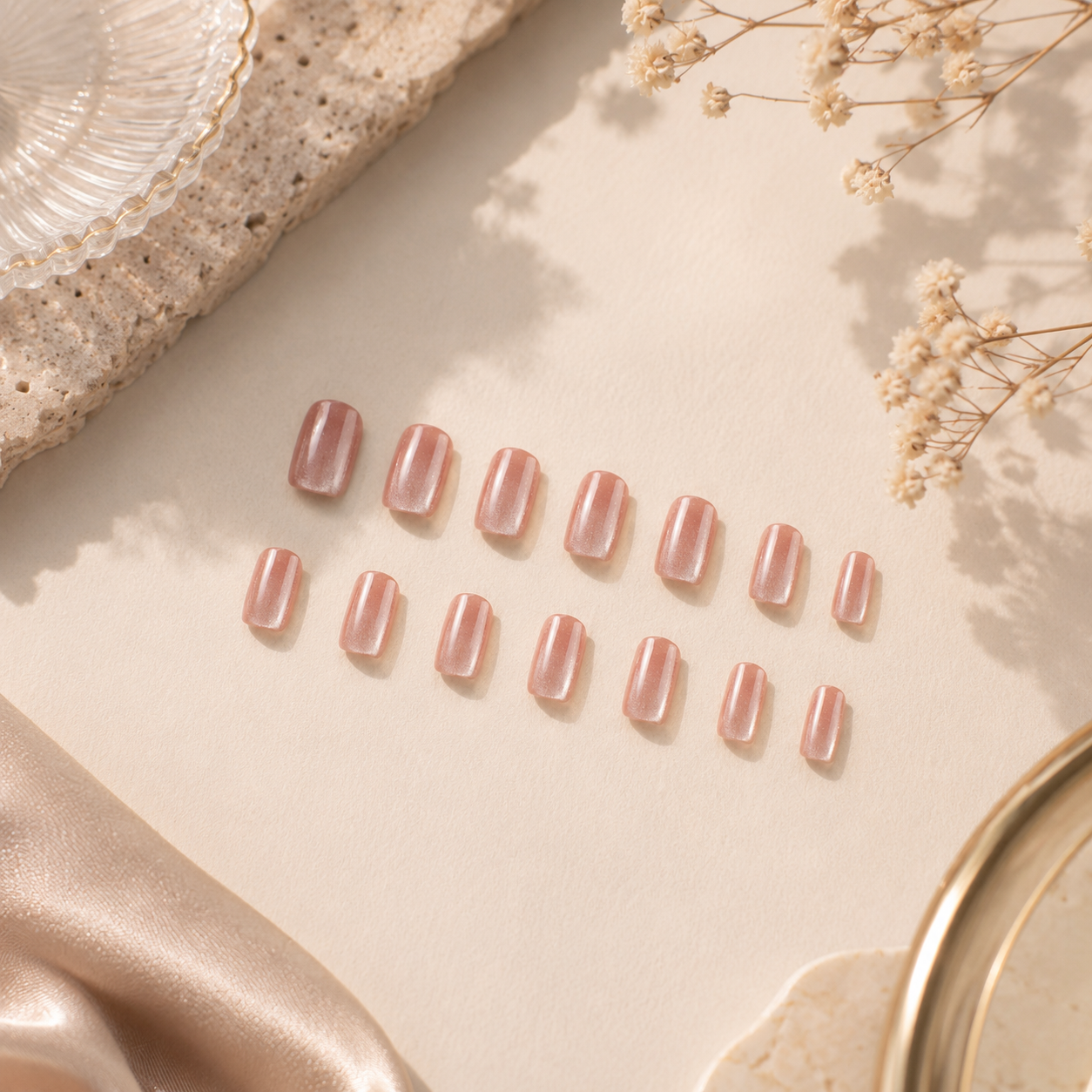 Set of glossy rose chrome press-on nails arranged on a cream background with dried flowers, textured stone decor, and soft natural lighting.